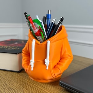 May include: An orange pencil holder shaped like a hooded sweatshirt with white drawstrings. The container is filled with various pens and markers in colors including red, blue, black, and green. The design features a front pocket and hood detail.