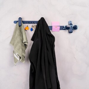 May include: A navy blue wall-mounted organizer with multiple hooks and a cross-shaped section. A green towel, keys, a black jacket, a pink sticky note, and sunglasses are hanging on the organizer. The wall is a light gray color.