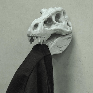 May include: A white, dinosaur skull-shaped wall hook. The skull has detailed features, including eye sockets and sharp teeth. A black garment hangs from the hook, contrasting with the skull's color. The hook is mounted on a light gray wall.