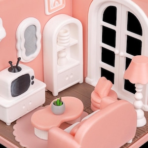 May include: A miniature pink and white dollhouse living room scene. The scene includes a sofa, armchair, coffee table, TV, lamp, and a decorative shelf. The walls are pink, and the furniture is a matching pink and white color scheme.