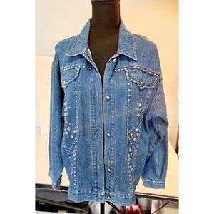 May include: A medium-wash denim jacket with silver-tone button closures and decorative rhinestone and turquoise-colored embellishments. The jacket has a classic collar and long sleeves. The embellishments are arranged in a pattern along the front and on the shoulders.