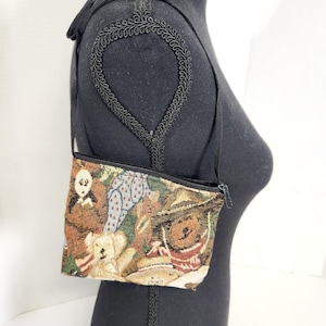 May include: A small tapestry shoulder bag featuring a repeating pattern of teddy bears in various poses and outfits. The bag has a black zipper and a thin black strap. The bag is displayed on a black mannequin.