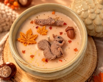 May include: A round ceramic bowl filled with light peach-colored wax, featuring embedded wax decorations including a brown squirrel, a textured log, a brown acorn, and several orange maple leaves, accented with small gold glitter flakes.