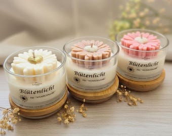 May include: Three flower-shaped candles in glass containers. One is white with a gold center, another is light brown, and the third is pink. Each candle sits on a small wooden coaster. The text "Blütenlicht" is printed on the glass.