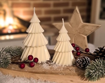 May include: Two ivory-colored, tiered Christmas tree candles, a star-shaped decoration, and red berries are arranged on a wooden surface. A pine cone and greenery add to the festive holiday decor. The background features a fireplace.
