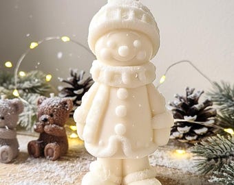 May include: A white snowman-shaped candle with a hat, scarf, and coat. The candle has a smiling face with button eyes and a carrot nose. The snowman is surrounded by pine cones, teddy bears, and Christmas lights.