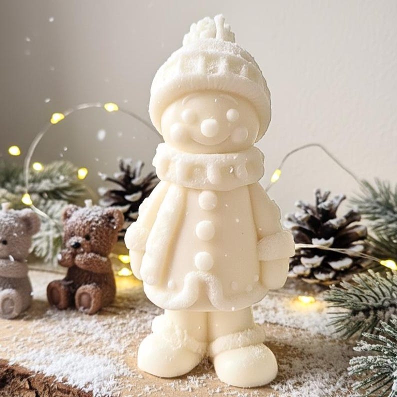 May include: A white snowman-shaped candle with a hat, scarf, and coat. The candle has a smiling face with button eyes and a carrot nose. The snowman is surrounded by pine cones, teddy bears, and Christmas lights.