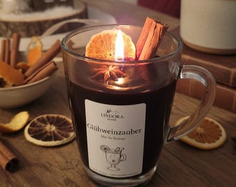 May include: A lit candle in a clear glass mug, filled with a dark liquid and garnished with a dried orange slice, cinnamon stick, and star anise. The mug has a handle and a label that reads "Glühweinzauuber". The candle sits on a wooden surface.