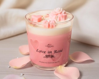 May include: A clear glass candle jar filled with cream-colored wax, topped with pink wax shapes including a flower and hearts. The label reads Lindora Love in Rosé natural soy wax handmade with love, surrounded by scattered pink rose petals.
