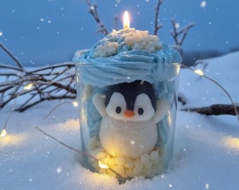 May include: A whimsical candle shaped like a penguin inside a clear glass. The penguin is white with a black head and orange beak, surrounded by blue frosting and topped with a lit candle. The candle sits in a snowy landscape with fairy lights.
