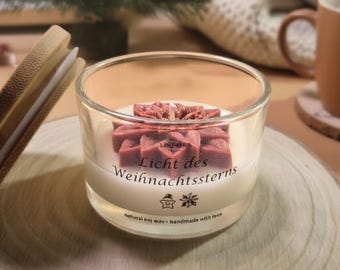May include: A clear glass candle holder with a white candle and a red star-shaped wax design. The candle has text that reads "Licht des Weihnachtssterns" and "natural soy wax - handmade with love". The candle sits on a wooden surface.