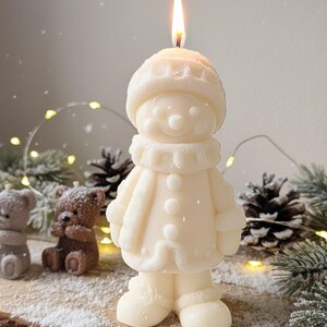 May include: A lit, ivory-colored snowman candle with a hat, scarf, and coat. The candle is standing on a wooden surface with other decorative items, including pine cones and small bear candles. The flame is bright.