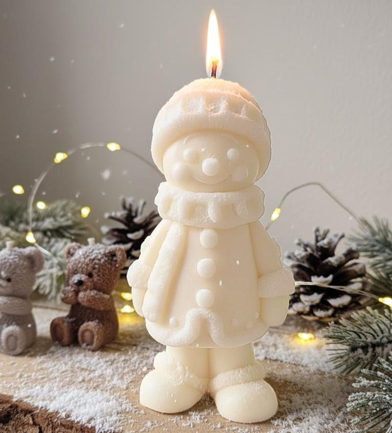 May include: A lit, ivory-colored snowman candle with a hat, scarf, and coat. The candle is standing on a wooden surface with other decorative items, including pine cones and small bear candles. The flame is bright.