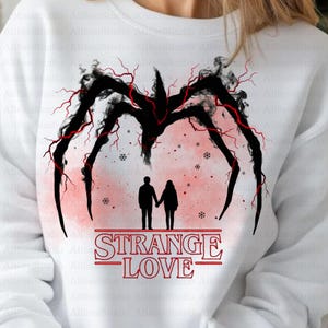 Puede incluir: Sudadera blanca con un diseño negro similar a una araña y detalles en rojo, con el texto "STRANGE LOVE" en rojo. La imagen incluye siluetas de una pareja tomadas de la mano y copos de nieve.