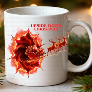 May include: White ceramic mug featuring a festive Christmas design. The mug displays Santa Claus in a red sleigh pulled by reindeer, emerging from a fiery portal. Text on the mug reads "UPSIDE DOWN CHRISTMAS".