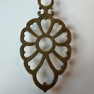 Puede incluir: Un salvamanteles decorativo de latón con un diseño floral. El salvamanteles presenta un círculo central rodeado de recortes en forma de corazón, con un asa ornamentada en la parte superior. El metal tiene un acabado cálido y antiguo.