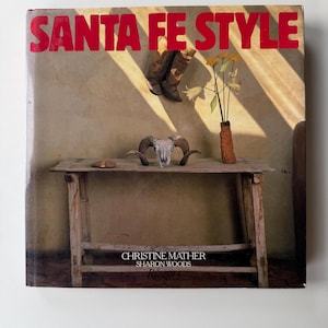 May include: A book cover titled "SANTA FE STYLE" in red letters. The cover features a rustic wooden table with a ram skull, a vase with yellow flowers, and a pair of cowboy boots. The authors' names are also visible.