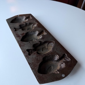 Puede incluir: Un molde de pescado vintage de hierro fundido con cinco impresiones detalladas de peces. El molde tiene forma rectangular con una pátina oscura y envejecida, y un pequeño agujero para colgar. Los diseños de peces presentan escamas y aletas texturizadas.