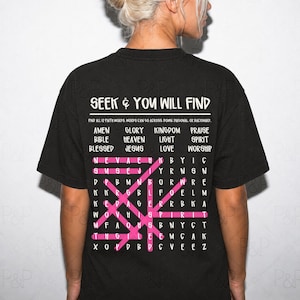 May include: A black t-shirt featuring a white word search puzzle graphic on the back. The text reads SEEK & YOU WILL FIND, followed by a list of 12 faith words and a grid of letters with several words highlighted by pink horizontal and diagonal lines.