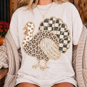 May include: A light beige short-sleeve t-shirt featuring a large graphic of a turkey. The turkey design incorporates various patterns including brown leopard print, black and white checkered squares, polka dots, and vertical stripes with gold outlines.