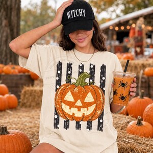 Peut inclure: Un t-shirt beige &agrave; manches courtes orn&eacute; d'un motif de citrouille jack-o'-lantern orange centr&eacute; sur des rayures verticales noires et blanches &agrave; pois blancs. Une casquette noire avec le texte blanc WITCHY est port&eacute;e sur la t&ecirc;te.