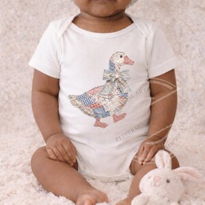 May include: White baby onesie featuring a patchwork goose design with a blue bow. The goose has a red, white, and blue patchwork pattern. The onesie is short-sleeved and has a simple, classic design.