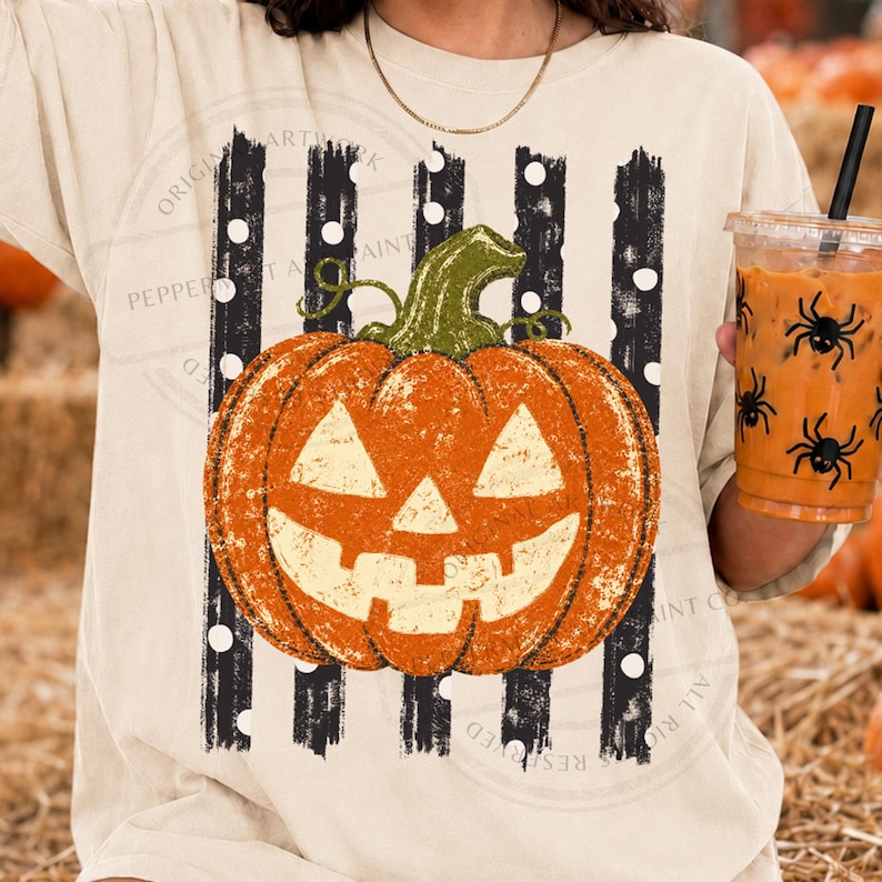 Peut inclure: Un t-shirt couleur cr&egrave;me arborant un motif de citrouille orange avec une tige verte, centr&eacute; sur des rayures verticales noires &agrave; pois blancs. La citrouille pr&eacute;sente une texture vieillie et un visage sculpt&eacute; de type jack-o'-lantern.
