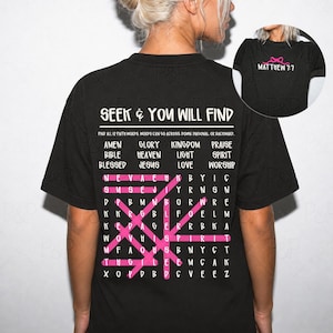 May include: A black t-shirt featuring a white word search puzzle on the back with the text SEEK AND YOU WILL FIND and a list of faith words. Pink lines highlight words within the grid. A small circular inset shows the front with MATTHEW 7:7 text.