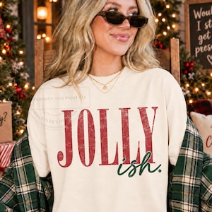 Può includere: Una felpa girocollo color crema con la scritta JOLLY in grandi lettere maiuscole rosse dall'aspetto vissuto, con la parola ish in un carattere corsivo verde scuro più piccolo sottostante. Il capo presenta una vestibilità rilassata.