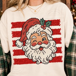 Könnte beinhalten: Ein cremefarbenes T-Shirt mit Rundhalsausschnitt und einer Grafik eines zwinkernden Weihnachtsmanns mit roter Mütze und Stechpalmenbeeren. Das Design hat eine strukturierte Strickoptik vor horizontalen roten Streifen. Der Text lautet Original Artwork Peppermint and Paint Co.