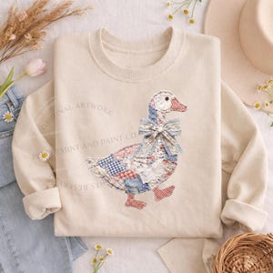 May include: Cream-colored sweatshirt featuring a patchwork goose design with red, blue, and white patterns. The goose has a blue bow and red feet. The text "ORIGINAL ARTWORK" and "PEPPERMINT AND PAINT CO." are printed on the sweatshirt.