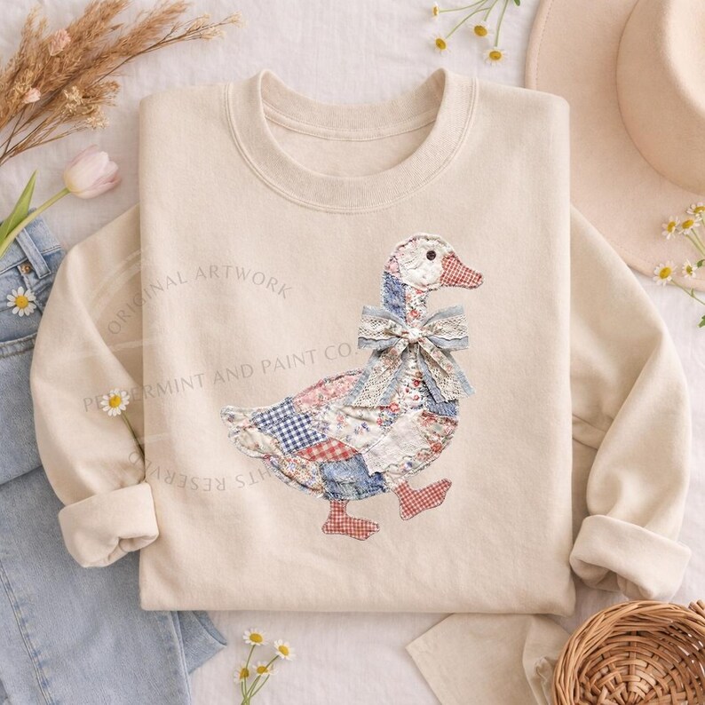 May include: Cream-colored sweatshirt featuring a patchwork goose design with red, blue, and white patterns. The goose has a blue bow and red feet. The text "ORIGINAL ARTWORK" and "PEPPERMINT AND PAINT CO." are printed on the sweatshirt.