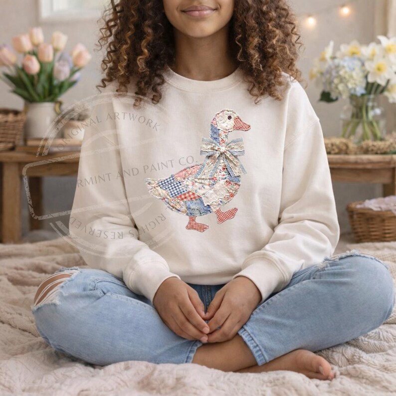 May include: Cream-colored sweatshirt featuring a patchwork goose design with red, blue, and white patterns. The goose has a bow and red feet. The sweatshirt is worn by a person sitting cross-legged.