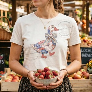 May include: Cream-colored t-shirt featuring a patchwork goose design with red, blue, and white patterns. The goose has a bow and red feet. The person is holding a bowl of fresh strawberries. The shirt has the text "ORIGINAL ARTWORK PERMINT AND PAINT CO."