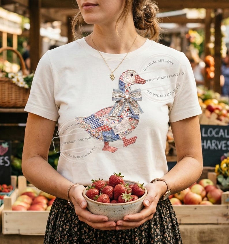 May include: Cream-colored t-shirt featuring a patchwork goose design with red, blue, and white patterns. The goose has a bow and red feet. The person is holding a bowl of fresh strawberries. The shirt has the text "ORIGINAL ARTWORK PERMINT AND PAINT CO."