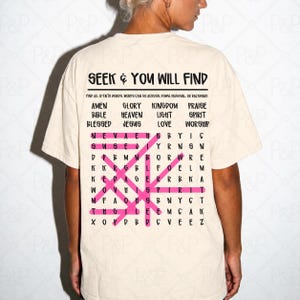 May include: A cream-colored short-sleeve t-shirt featuring a black word search puzzle graphic on the back. The text reads SEEK & YOU WILL FIND, followed by a list of faith words and a grid with several words highlighted in bright pink.