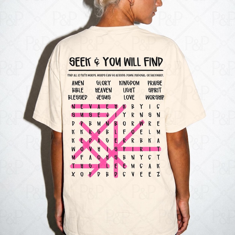 May include: A cream-colored short-sleeve t-shirt featuring a black word search puzzle graphic on the back. The text reads SEEK & YOU WILL FIND, followed by a list of faith words and a grid with several words highlighted in bright pink.