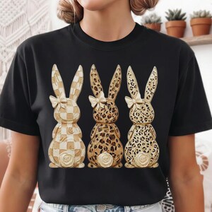 May include: Black t-shirt featuring three decorative bunny figures. The bunnies have different patterns: checkered, leopard print, and dotted. Each bunny has a white bow and a rose detail at the base. The shirt is made of a soft material.