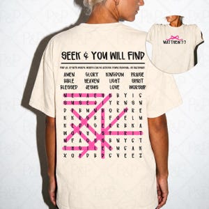 May include: A cream-colored t-shirt featuring a word search puzzle on the back with the text SEEK & YOU WILL FIND and a list of faith-based words. Several words are highlighted with pink lines. The front chest area displays a small pink bow and the text MATTHEW 7:7.