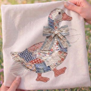 May include: A decorative, patchwork goose design on a cream-colored fabric. The goose features a patchwork of floral, gingham, and solid fabrics in shades of red, blue, and cream. A blue bow with lace trim adorns the neck. The text "PEPPERMINT AND PAINT CO." is visible.