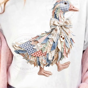 May include: White t-shirt featuring a whimsical goose design. The goose is adorned with fabric scraps in various patterns, including gingham and floral prints. A large bow accents the neck. The artwork has a patchwork aesthetic, with a mix of colors and textures.