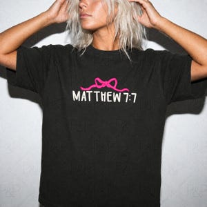 May include: A black short-sleeved t-shirt featuring a graphic design on the chest. The design includes a bright pink bow icon positioned above the white text that reads MATTHEW 7:7 in a clean, sans-serif font. The shirt has a crew neckline.