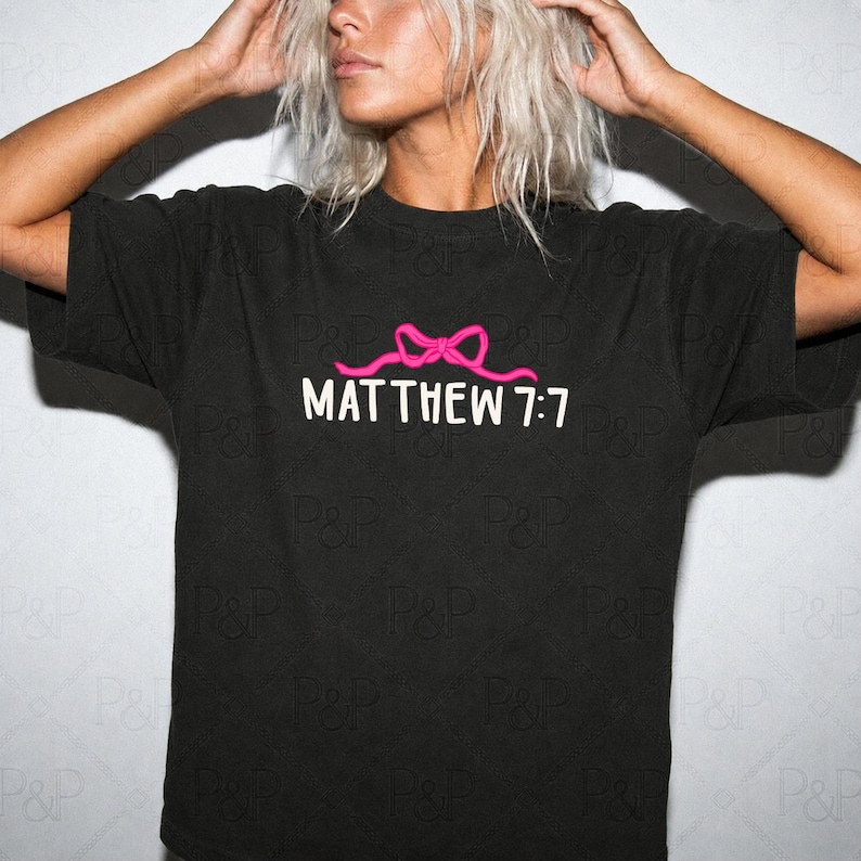 May include: A black short-sleeved t-shirt featuring a graphic design on the chest. The design includes a bright pink bow icon positioned above the white text that reads MATTHEW 7:7 in a clean, sans-serif font. The shirt has a crew neckline.