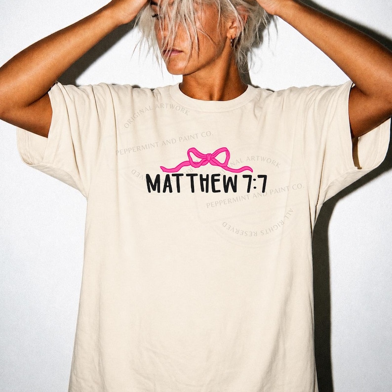 May include: A beige short-sleeved t-shirt featuring a graphic design on the chest. The design includes a bright pink ribbon bow positioned above the black text that reads MATTHEW 7:7. The shirt has a crew neckline and a relaxed, loose-fitting cut.