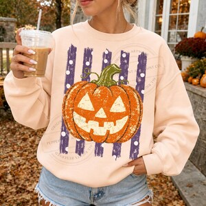 Peut inclure: Un sweat-shirt &agrave; col rond couleur p&ecirc;che clair orn&eacute; d'un motif de citrouille orange sculpt&eacute;e en jack-o'-lantern, centr&eacute; sur des rayures verticales violettes &agrave; pois blancs. La citrouille a une tige verte et une texture vieillie.