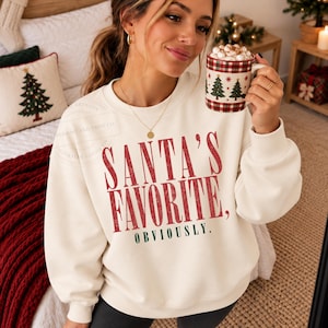 Puede incluir: Una sudadera de cuello redondo color crema con el texto en rojo "SANTA'S FAVORITE," y el texto en verde "OBVIOUSLY." debajo. La sudadera tiene mangas largas y cuenta con cuello, puños y dobladillo acanalados para un ajuste cómodo.