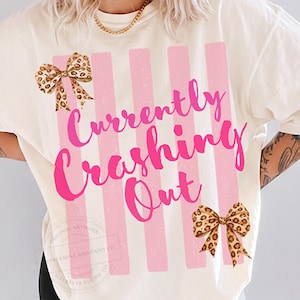 May include: Cream-colored t-shirt with pink vertical stripes and the phrase "Currently Crashing Out" in a vibrant pink script. The shirt features two leopard print bows, one at the top left and one at the bottom right.