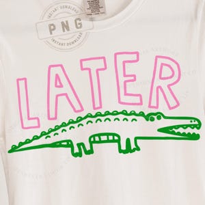 May include: White t-shirt with the word "LATER" in pink block letters above a green alligator illustration. The design is simple and bold, perfect for casual wear.