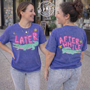 May include: Two women wearing blue t-shirts with alligator graphics. The front of the shirt says "LATER" with an alligator design. The back of the shirt says "AFTER WHILE" with a matching alligator. The shirts are paired with light wash jeans.