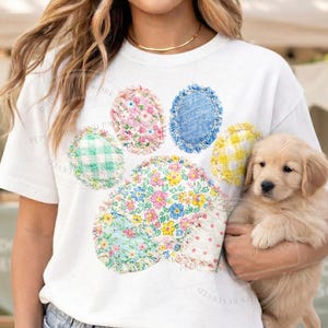 Puede incluir: Camiseta blanca con un diseño de huella de pata colorida hecha de parches de tela floral y estampada. Se sostiene un cachorro de golden retriever. La camiseta tiene las palabras "ORIGINAL PEPPER WORK AND PA" impresas.